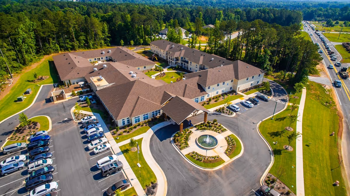 Aerial view of a senior living facility with a large building, circular driveway and fountain, parking lots, and surrounding trees.