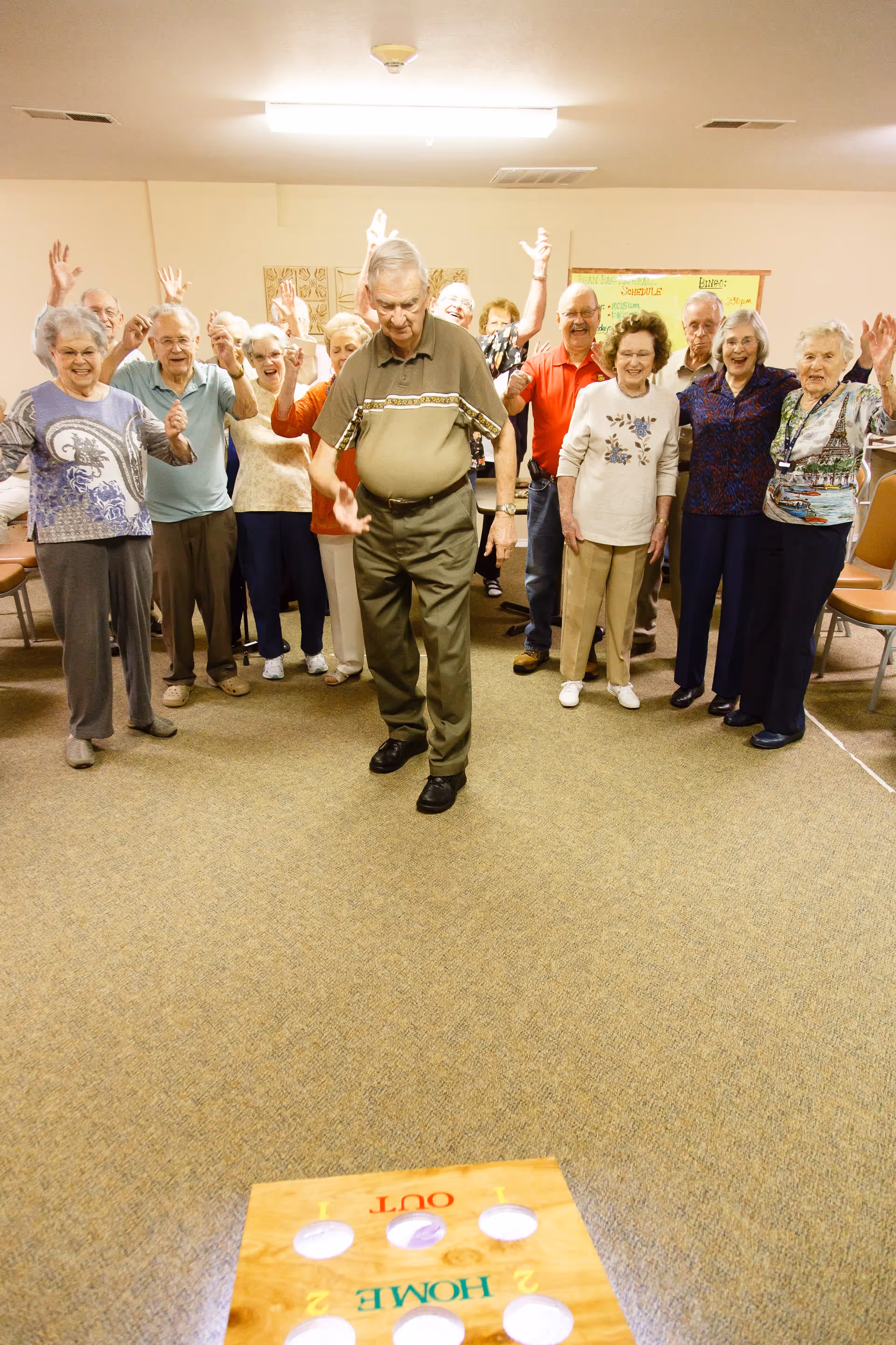 A group of elderly people standing in a room, enthusiastically watching and cheering as an elderly man in the front prepares to toss a bean bag into a wooden cornhole board on the floor. The room has beige walls and carpet, with chairs along the sides.
