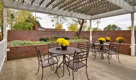 Outdoor patio area with metal tables and chairs under a pergola. Each table has a pot of yellow flowers. There is a brick wall surrounding the patio with some greenery and trees visible beyond it.