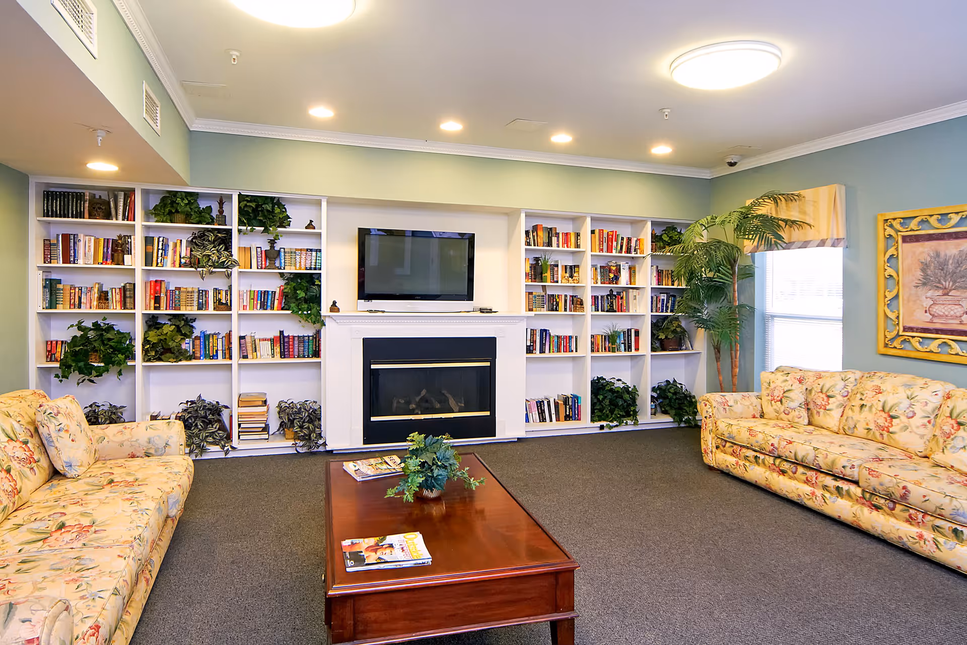 A cozy living room with two floral-patterned sofas facing a wooden coffee table. Behind the table is a white fireplace with a flat-screen TV mounted above it. Surrounding the fireplace are white built-in bookshelves filled with books and green plants. A large window with a yellow valance and a framed artwork are on the right wall. The room has soft green walls and recessed ceiling lights.
