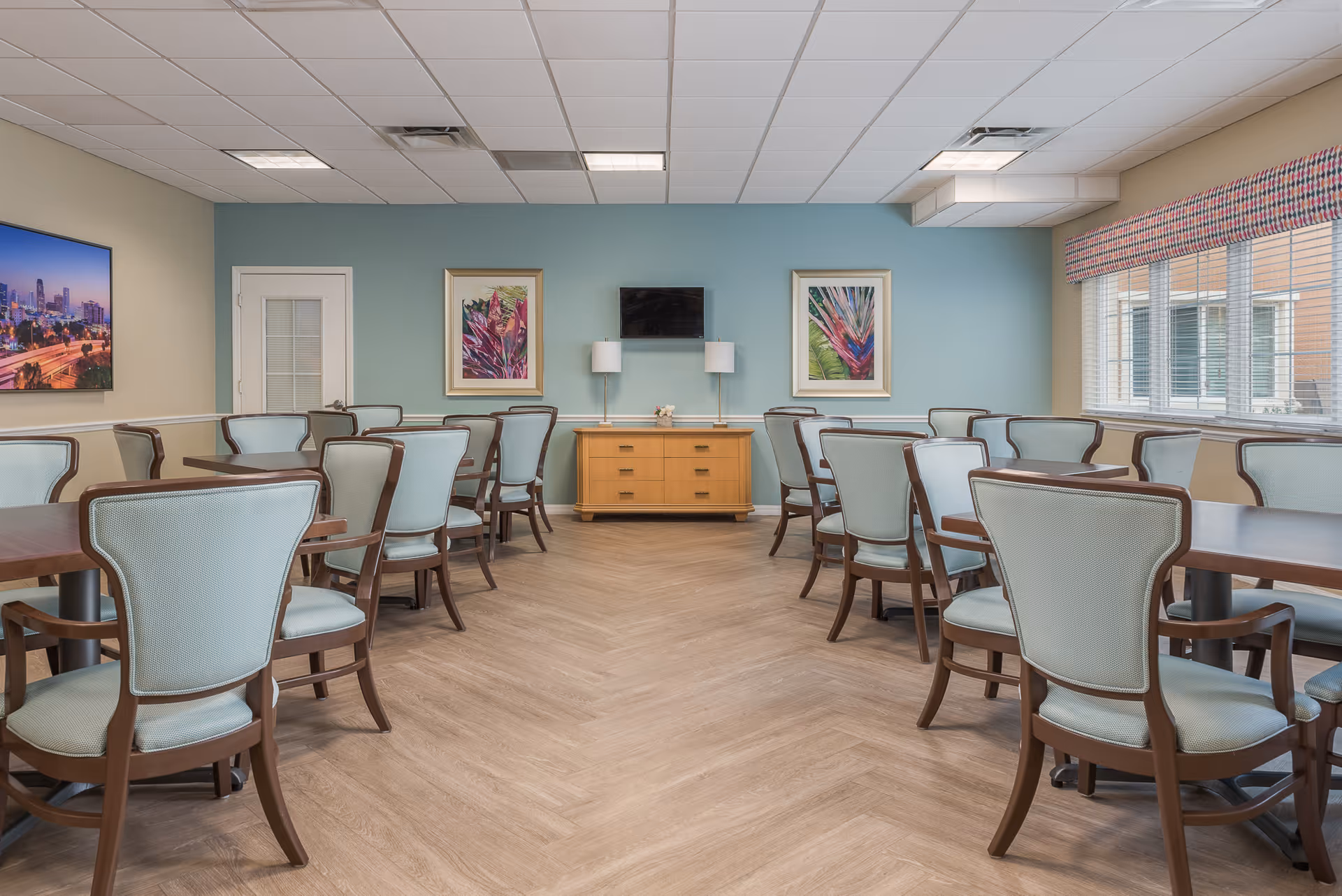 A dining room with multiple wooden tables and light blue cushioned chairs arranged neatly. The room has light wood flooring, a blue accent wall with two framed botanical prints, a flat-screen TV mounted on the wall, and two table lamps on a wooden sideboard. Large windows with blinds and a patterned valance allow natural light to enter the space.