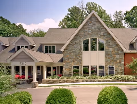Exterior view of Byron House, a two-story building with a stone facade and large windows. The entrance features a covered porch with white columns and the number 9210 displayed above. The surrounding area includes green bushes and trees under a clear sky.