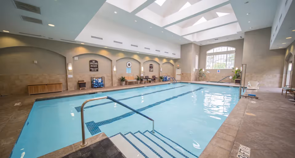 Indoor swimming pool with clear blue water, metal handrails, and steps leading into the pool. The pool area has beige tiled floors and walls, arched wall niches with benches and plants, large windows letting in natural light, and skylights in the ceiling.