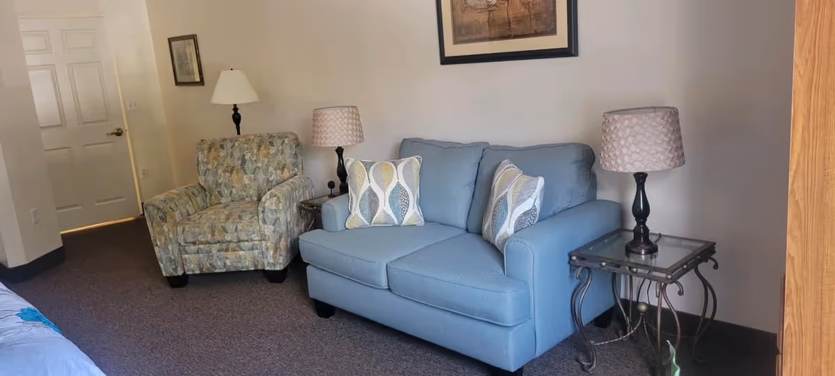 A light-blue loveseat and patterned armchair in a furnished living area with side tables, lamps, and wall art.