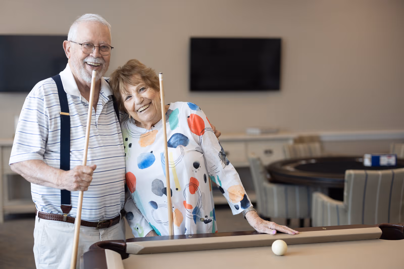 An elderly man and woman smiling and posing together while holding pool cues next to a pool table in a recreational room with a TV and a round table with chairs in the background.