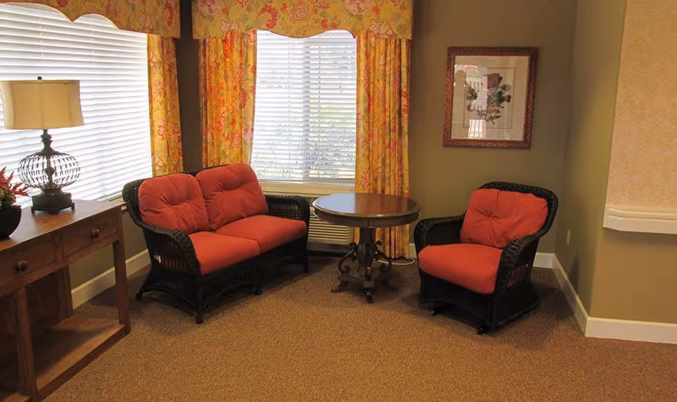 A cozy sitting area with a small round wooden table, a cushioned loveseat, and a cushioned armchair with red cushions. The area is next to a window with yellow floral curtains and blinds. A wooden side table with a decorative lamp and a flower arrangement is also visible.