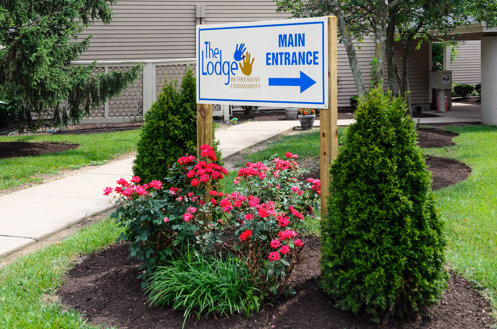A landscaped garden area with green shrubs and blooming red flowers surrounding a wooden post sign. The sign reads 'The Lodge Retirement Community MAIN ENTRANCE' with a blue arrow pointing to the right. A sidewalk and part of a building are visible in the background.