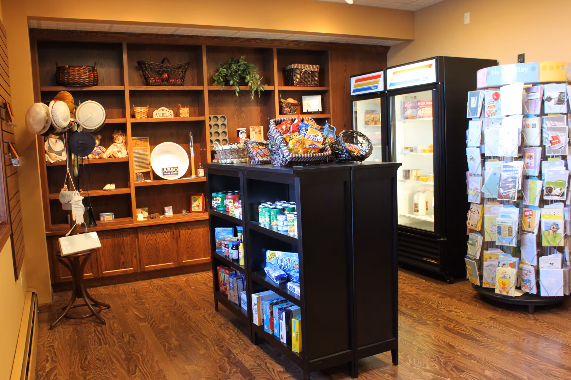 Interior view of a small shop area in a senior living facility with wooden shelves displaying hats, baskets, and decorative items. A central black shelving unit holds various snacks and canned goods. Two refrigerators with glass doors contain dairy and frozen products. A rotating rack displays greeting cards on the right side. The floor is wooden and the walls are painted warm tones.