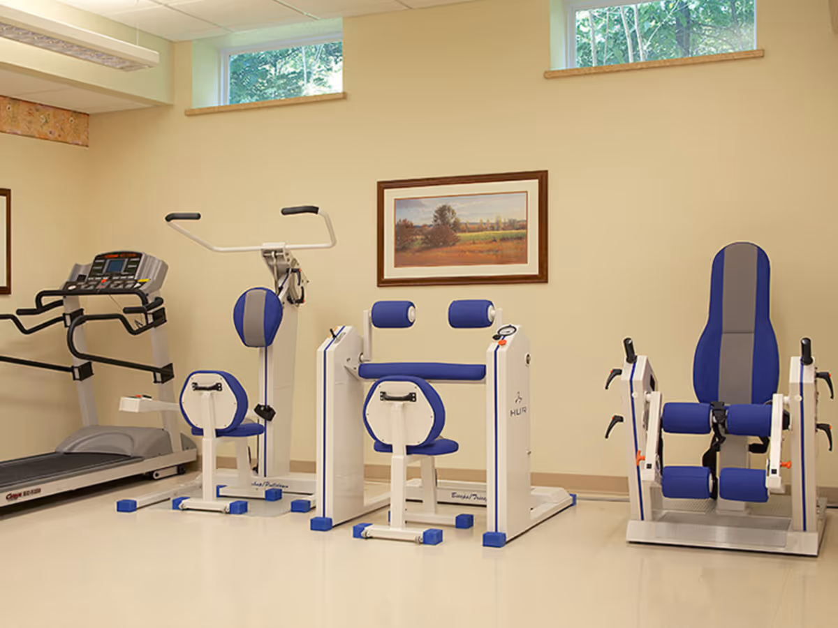 A small exercise room with various fitness equipment including a treadmill, a seated arm exercise machine, and a leg press machine. The walls are beige with two small windows near the ceiling and a framed landscape painting hanging on the wall.