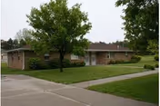 Single-story brick building with a brown roof surrounded by green grass and trees, with a concrete sidewalk and driveway in the foreground.