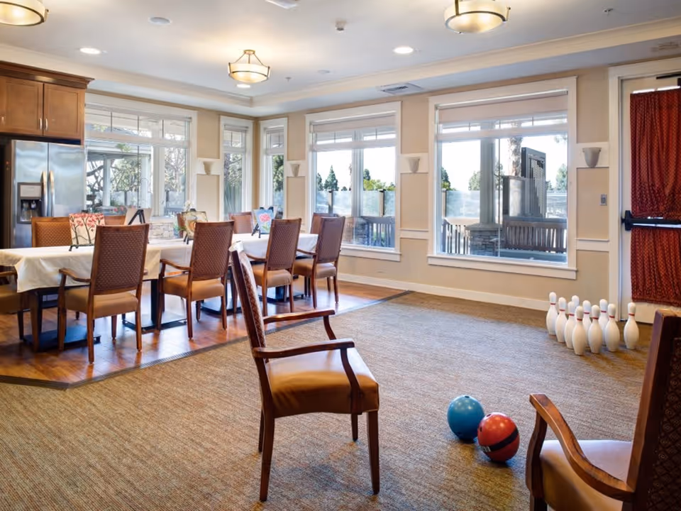 A bright and spacious room with large windows letting in natural light. The room features a dining area with a long table covered with a white tablecloth and surrounded by wooden chairs with cushioned seats. In the foreground, there are two chairs facing a small bowling setup with pins and two bowling balls on a carpeted floor. The room has wooden flooring near the dining area and carpet in the bowling area, with ceiling lights providing additional illumination.