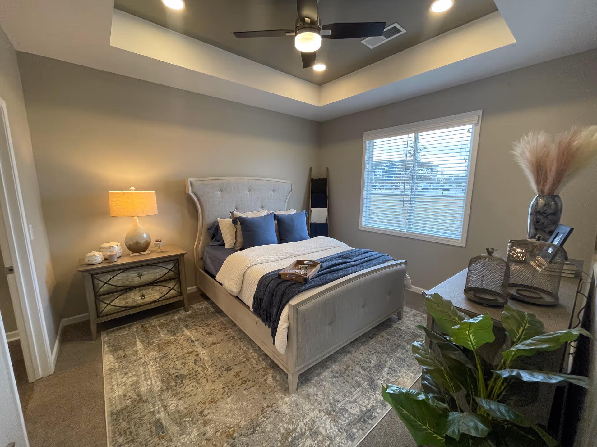 Bright, styled bedroom featuring a bed with navy pillows and throw, a nightstand with lamp, dresser, window, ceiling fan, and a potted plant.