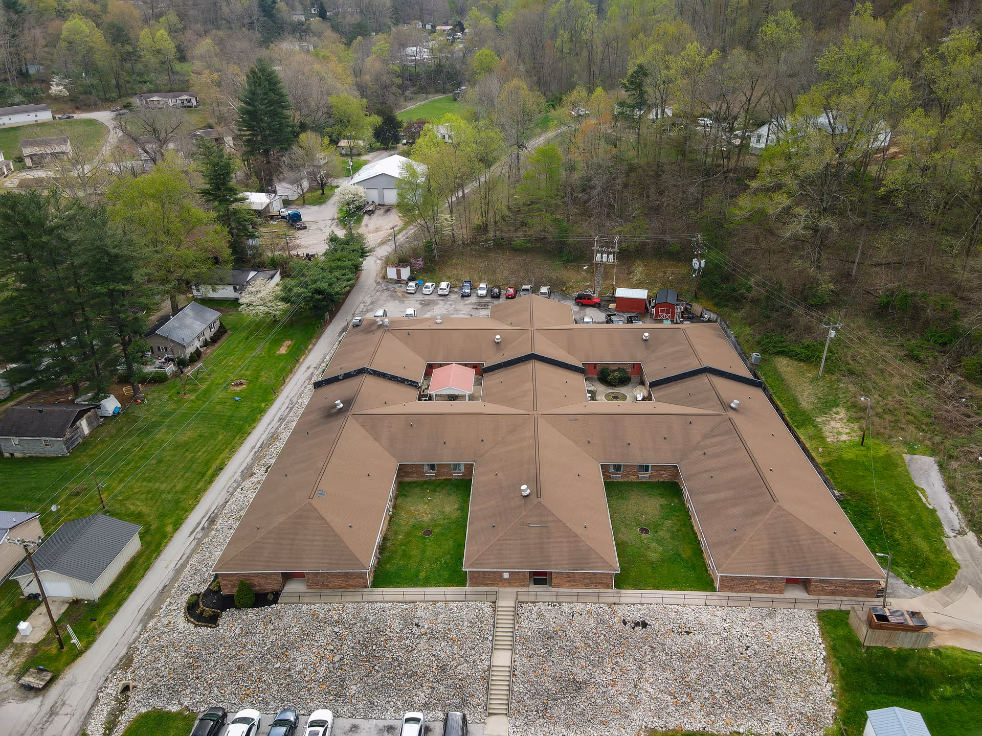 Aerial view of Vanceburg Hills facility showing a large, single-story building with a brown roof and multiple wings forming a U-shape. Surrounding the building are green lawns, trees, a parking lot with several cars, and nearby residential houses. The area is lush with greenery and some wooded hills in the background.
