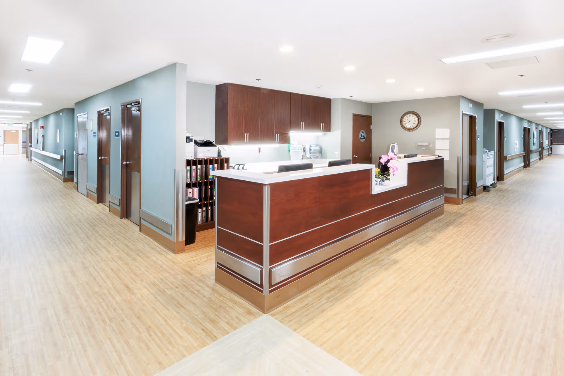 A bright, clean nursing station in a senior living facility hallway with wood flooring and light-colored walls. The nursing station has a dark wood counter with white trim, office chairs, and cabinets behind it. The hallway extends in both directions with multiple doors and handrails along the walls.