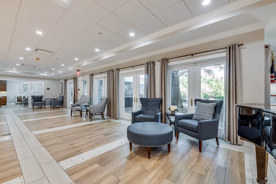 A spacious and well-lit senior living facility common area with multiple seating arrangements including gray armchairs and a round ottoman. Large windows with beige curtains allow natural light to fill the room, and a piano is visible on the right side. The floor has a wood and tile pattern, and the ceiling has recessed lighting.