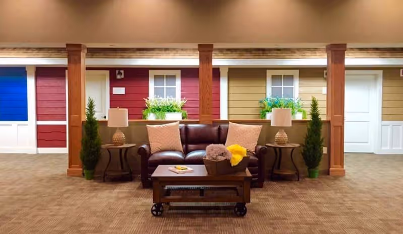 A cozy seating area with a dark brown leather sofa adorned with two light-colored pillows, flanked by two side tables each holding a lamp. In front of the sofa is a wooden coffee table with a basket containing folded blankets and a book. The background features a wall designed to look like the exterior of cottages with colorful siding in blue, red, and beige, white doors, and windows with flower boxes. There are also wooden columns and potted plants on either side of the sofa.