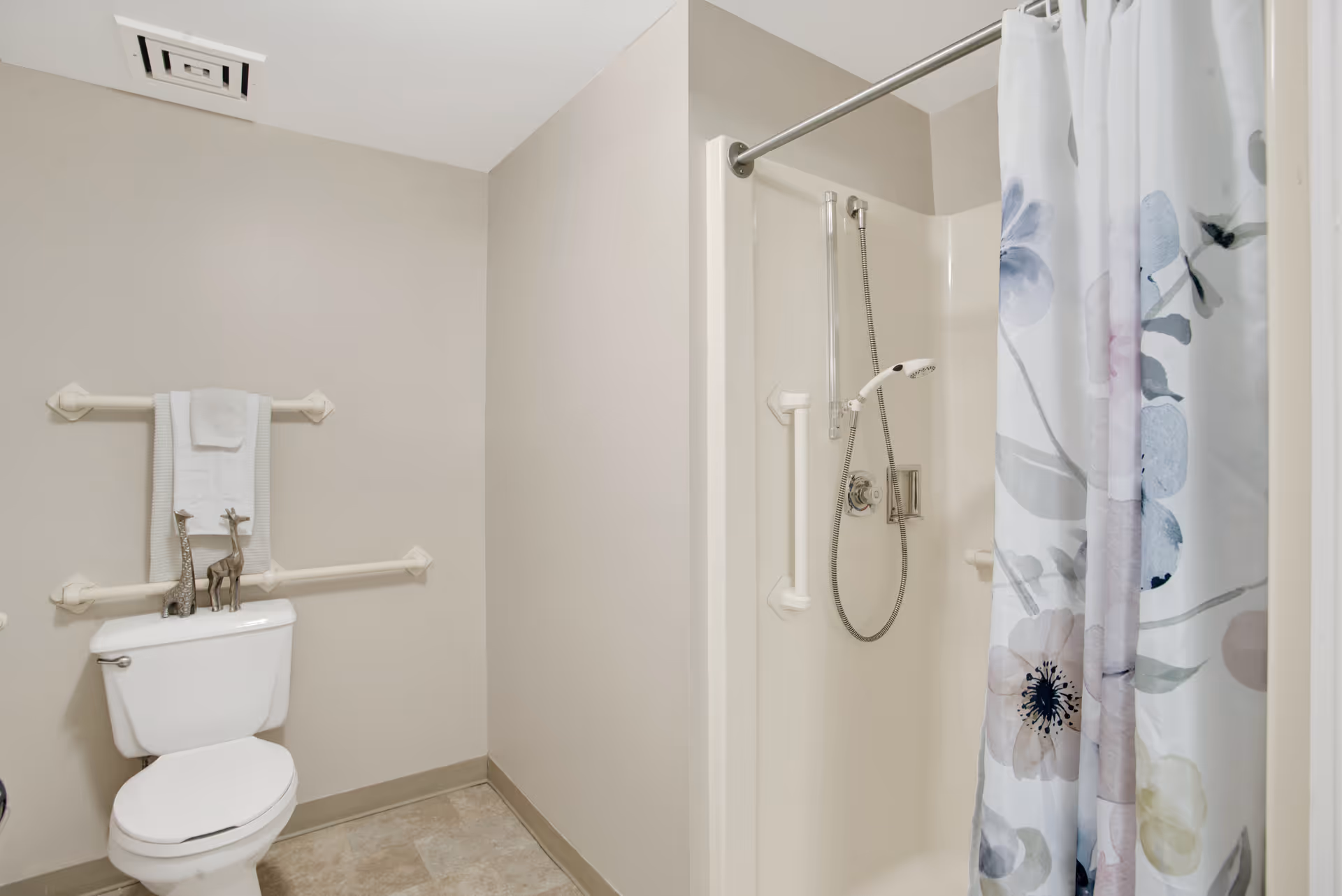 Accessible bathroom with a toilet and grab bars, towel on a rail, and a shower stall with a floral curtain.