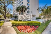 Exterior view of Hillcrest Hampton House, a multi-story residential building surrounded by trees and landscaping, with a sign in front displaying the facility's name and logo, and a flower bed with red flowers in the foreground.