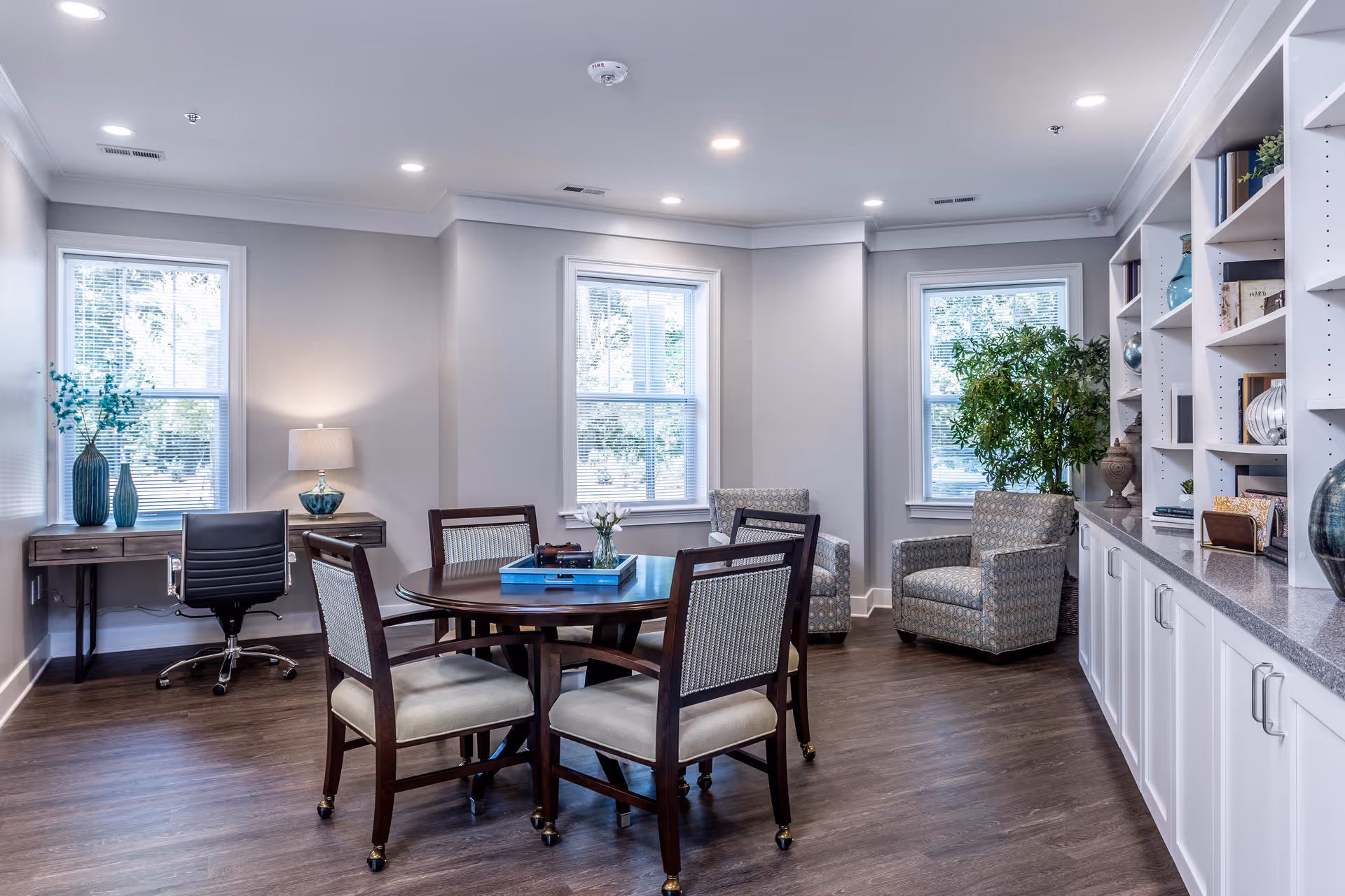 Bright common room with a round table and chairs, armchairs, built-in shelving, and a desk by the windows.