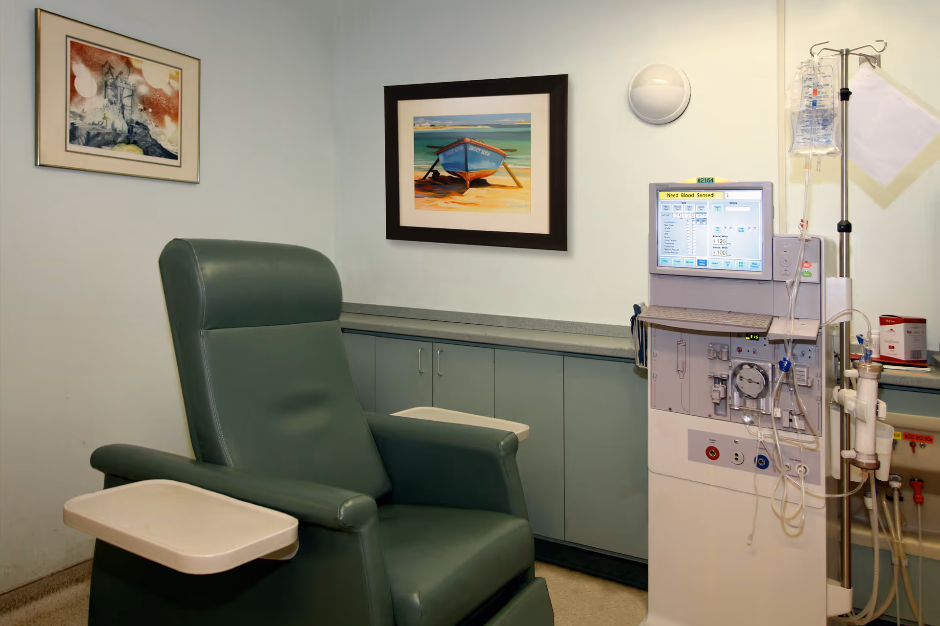 A medical treatment room with a green reclining chair featuring an attached tray, a dialysis machine with tubes and IV bags, light green cabinets along the wall, and two framed pictures hanging above the cabinets.