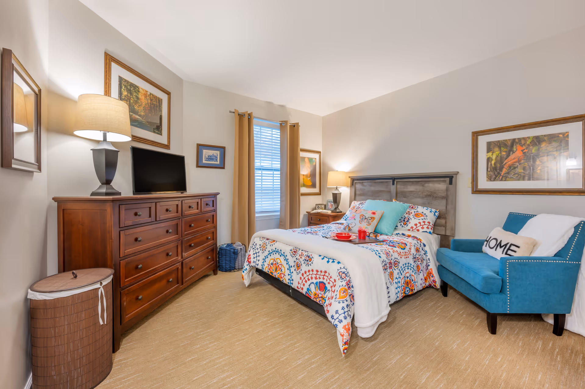 Well-lit bedroom with a bed dressed in colorful bedding, a wooden dresser with a lamp and TV, and a teal armchair.