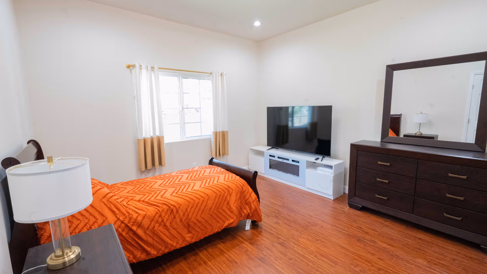 A bedroom with a single bed covered in an orange textured blanket, a bedside table with a lamp, a window with white and beige curtains, a large flat-screen TV on a white TV stand, and a dark wooden dresser with a large mirror above it. The room has wooden flooring and white walls.
