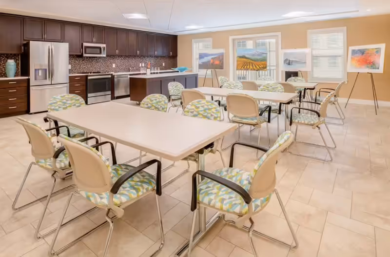 Bright communal dining room with multiple tables, cushioned chairs, and a kitchenette along back wall.
