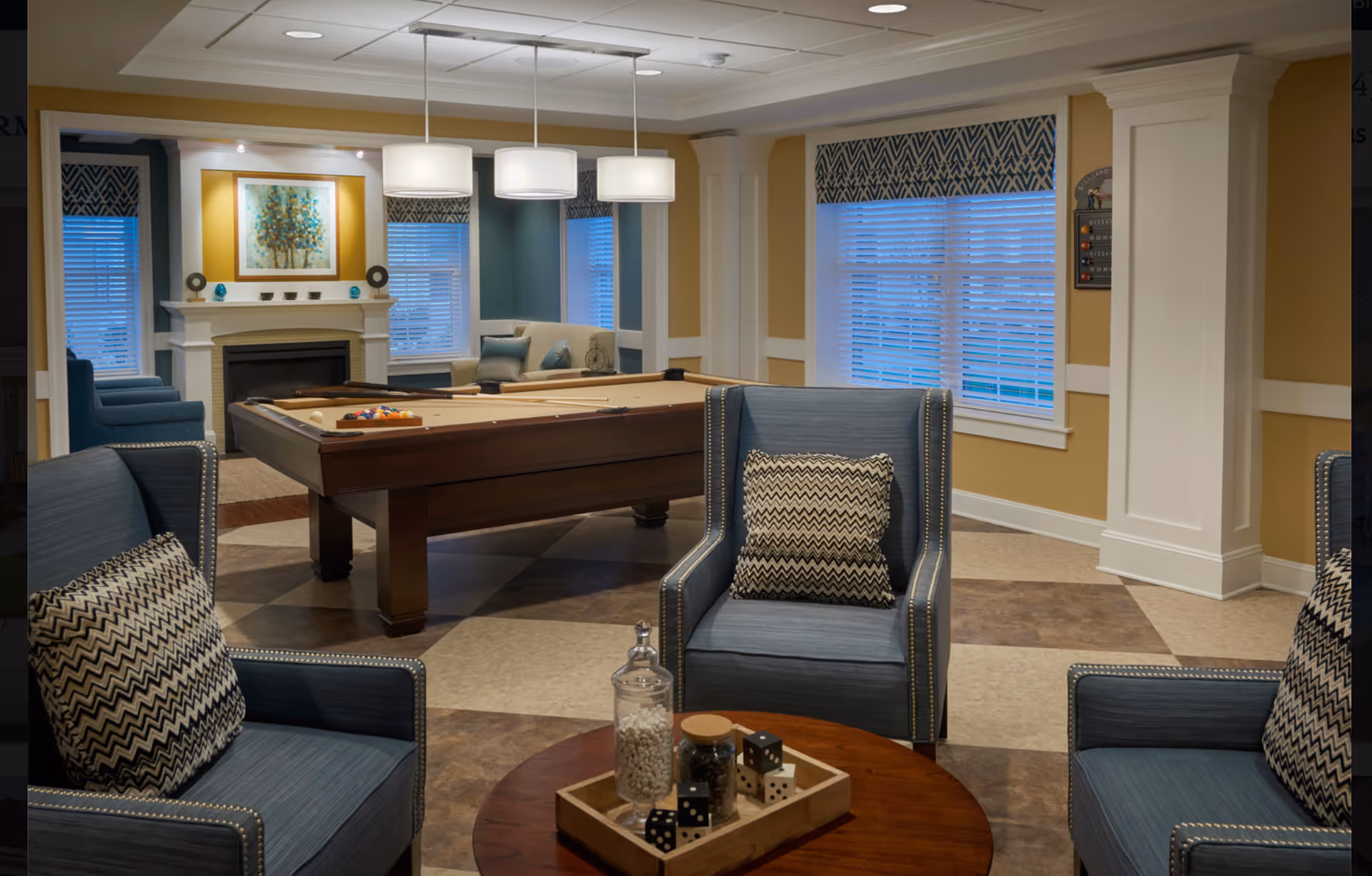 A cozy and well-lit common area featuring a pool table in the center, surrounded by comfortable blue armchairs with patterned cushions. The room has large windows with patterned valances, a fireplace with decorative items on the mantel, and a round wooden table with jars and dice as decor.