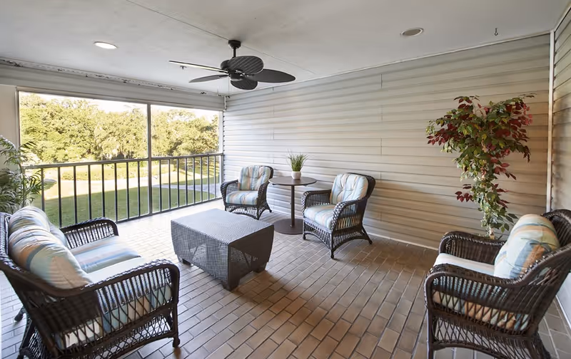 A covered outdoor patio area with wicker furniture including a sofa, four armchairs with cushions, a rectangular coffee table, and a small round table with a potted plant. The patio has a ceiling fan and overlooks a green lawn and trees through a railing.