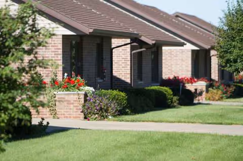 Brick senior living facility with front porches, manicured lawn, and colorful flowers along walkway.
