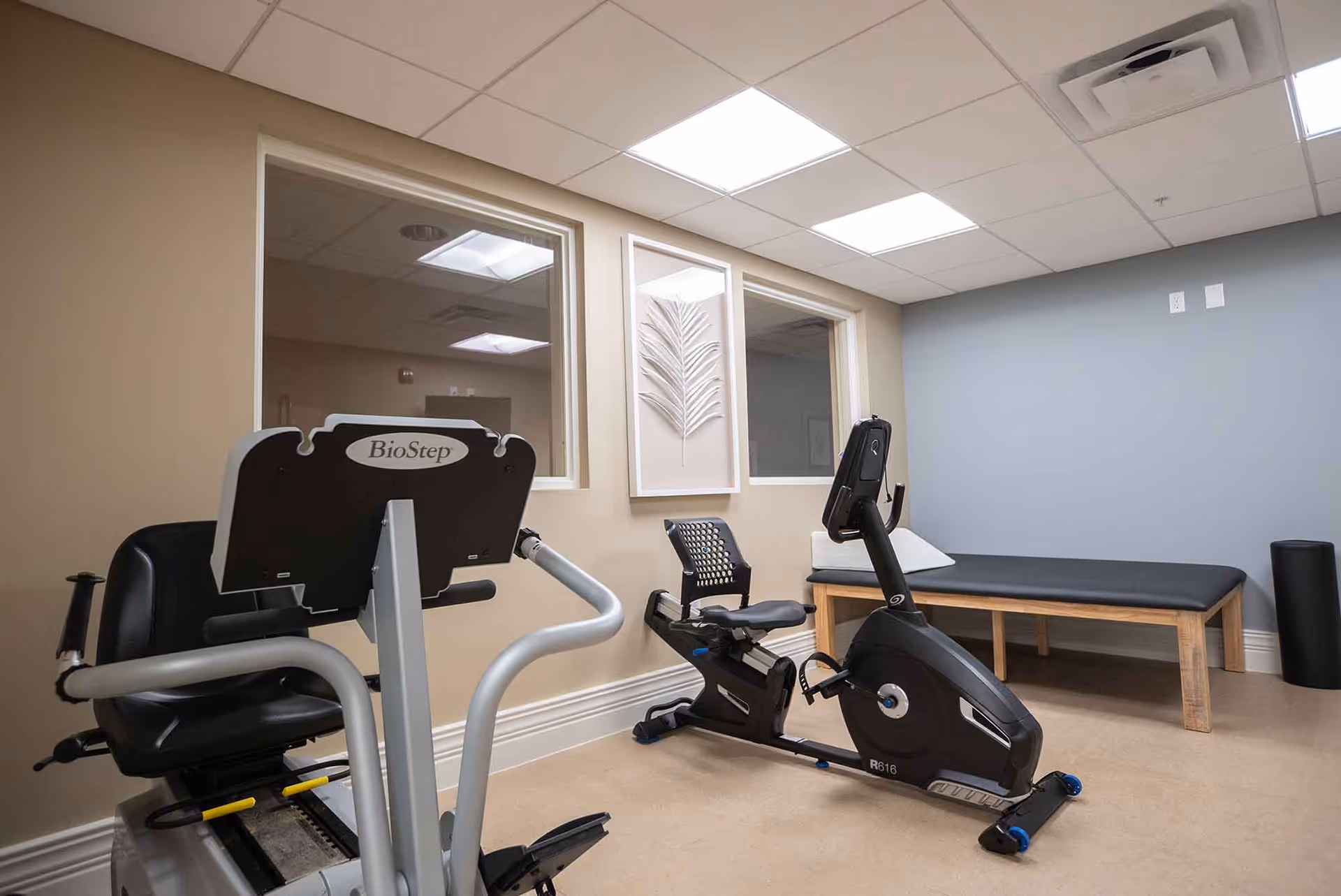 A small exercise room with two stationary exercise bikes, a padded bench against the wall, and a foam roller in the corner. The walls are painted beige and light blue, with two windows and a framed decorative piece between them. The ceiling has recessed lighting panels.