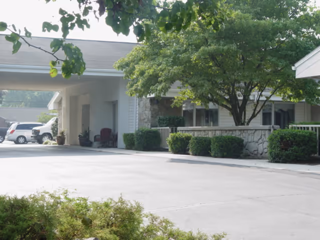Exterior view of a single-story building with a covered entrance, surrounded by green bushes and a large tree, with parked cars visible in the background.