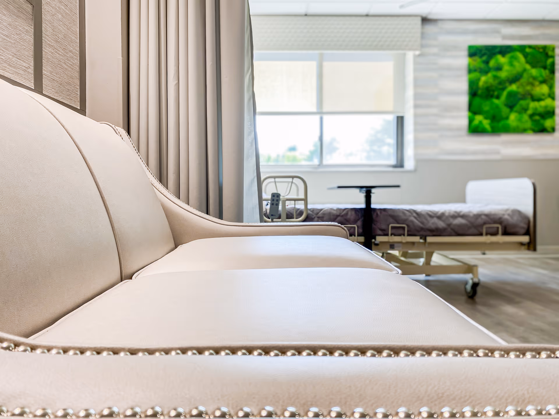 A close-up view of a beige leather sofa with nailhead trim in a well-lit room. In the background, there is a hospital-style bed with a quilted blanket, a window with blinds, and a green moss wall art piece mounted on a light-colored wall.