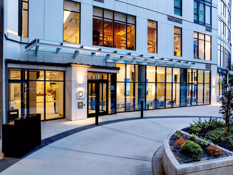 Exterior view of The Watermark At Bellevue Assisted Living & Memory Care building entrance at dusk, featuring large glass windows, a curved driveway, and landscaped plants in the foreground.