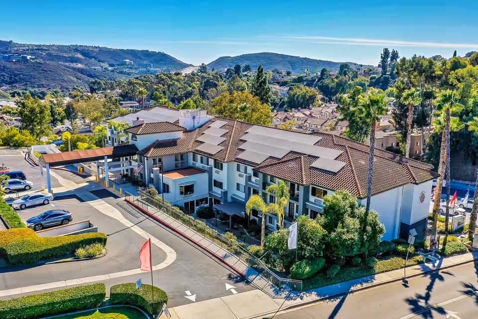 Aerial view of Rancho Penasquitos Senior Living facility showing a large two-story building with a tiled roof equipped with solar panels. The building is surrounded by trees, shrubs, and a parking lot with several cars. Hills and residential houses are visible in the background under a clear blue sky.