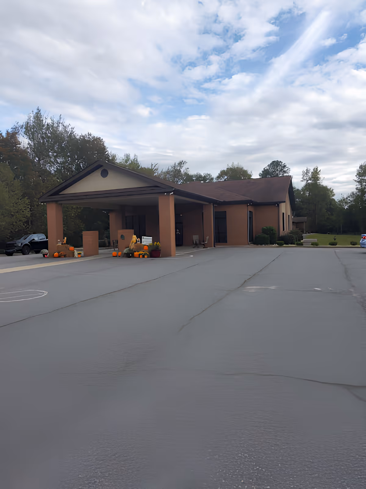 Exterior view of a single-story building with a covered entrance supported by four columns. The building is surrounded by trees and has a parking lot in front. There are fall decorations including pumpkins near the entrance.