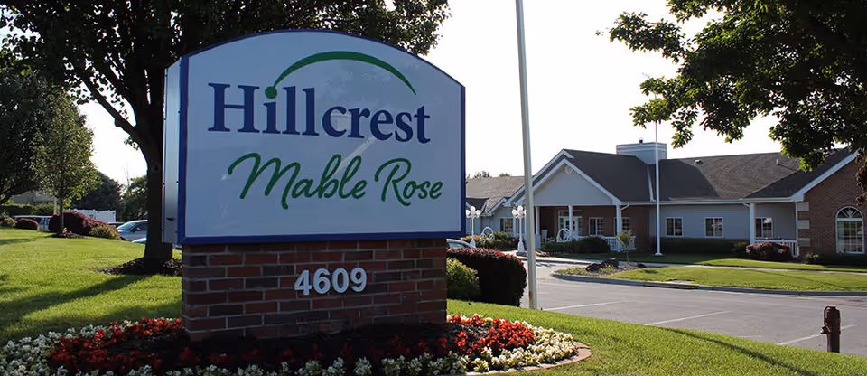 Outdoor view of the Hillcrest Mable Rose facility sign with the building and parking area in the background, surrounded by green grass, trees, and flower beds.