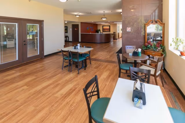 Bright dining area with several small tables and chairs on wood flooring and a reception desk visible in the background.