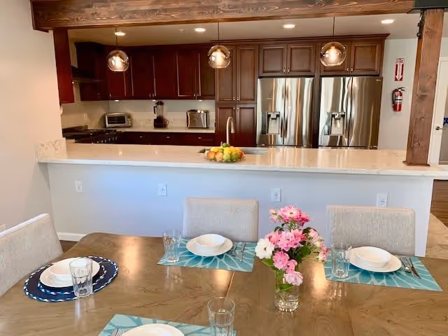 A dining table set with plates, glasses, and silverware, with a vase of pink and white flowers in the center. Behind the table is a kitchen with dark wood cabinets, stainless steel refrigerator, and three pendant lights hanging above a white countertop with a bowl of fruit.