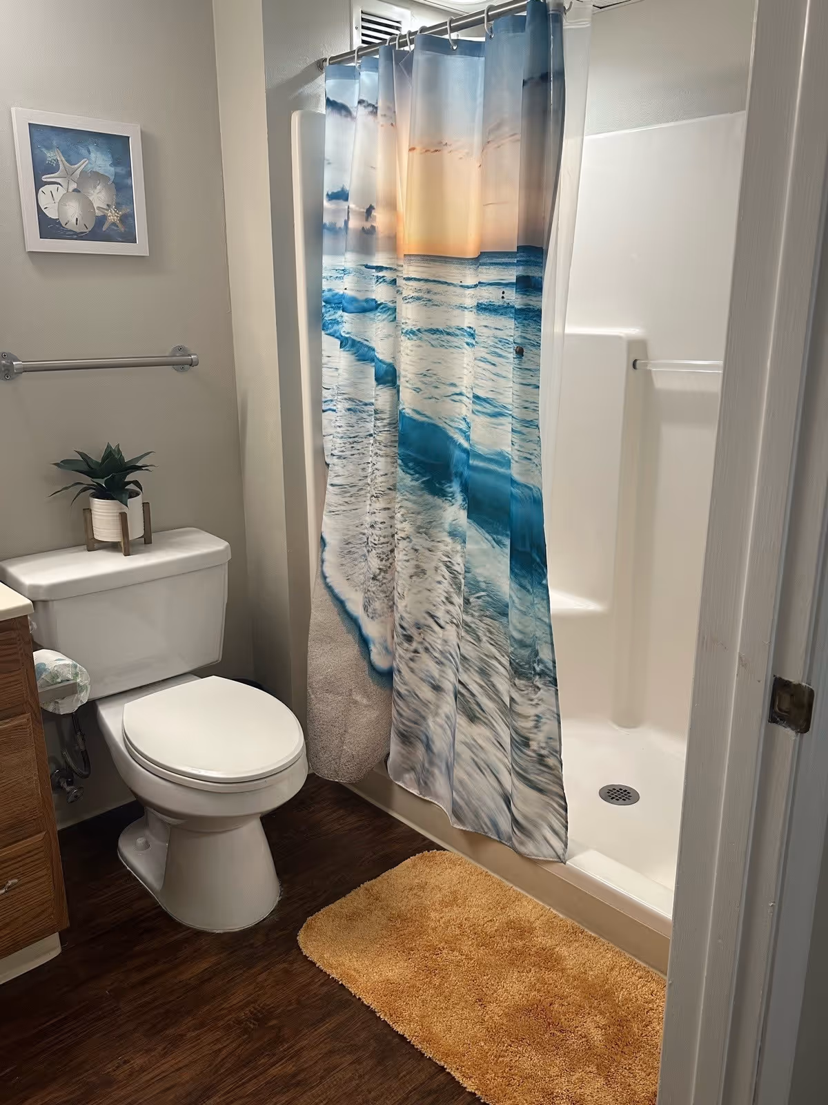 A bathroom with a white toilet, a wooden vanity partially visible on the left, and a shower with a curtain depicting a beach scene with waves and a sunset. There is a beige bath mat on the dark wood floor, a small potted plant on the toilet tank, a towel bar without a towel, and a framed picture of seashells on the wall.
