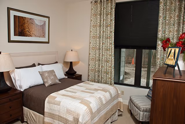 A cozy bedroom with a neatly made bed featuring a beige and brown quilt, white and brown pillows, two bedside tables each with a lamp, patterned curtains covering a window, a striped upholstered chair, a wooden dresser with a framed picture and a vase of red flowers, and a framed abstract artwork on the wall.
