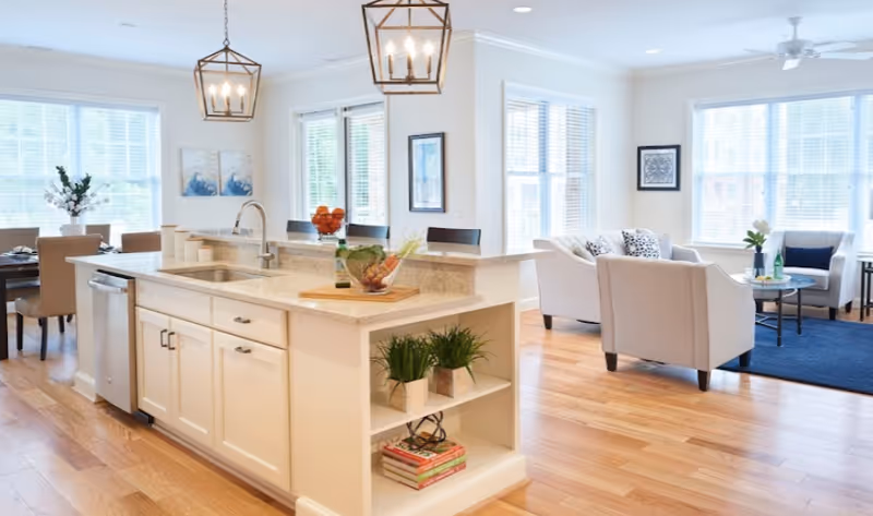Bright and spacious open-concept living area featuring a kitchen island with a sink and modern light fixtures, a dining area with a table and chairs near large windows, and a living room with light-colored sofas, armchairs, a blue rug, and framed artwork on the walls.