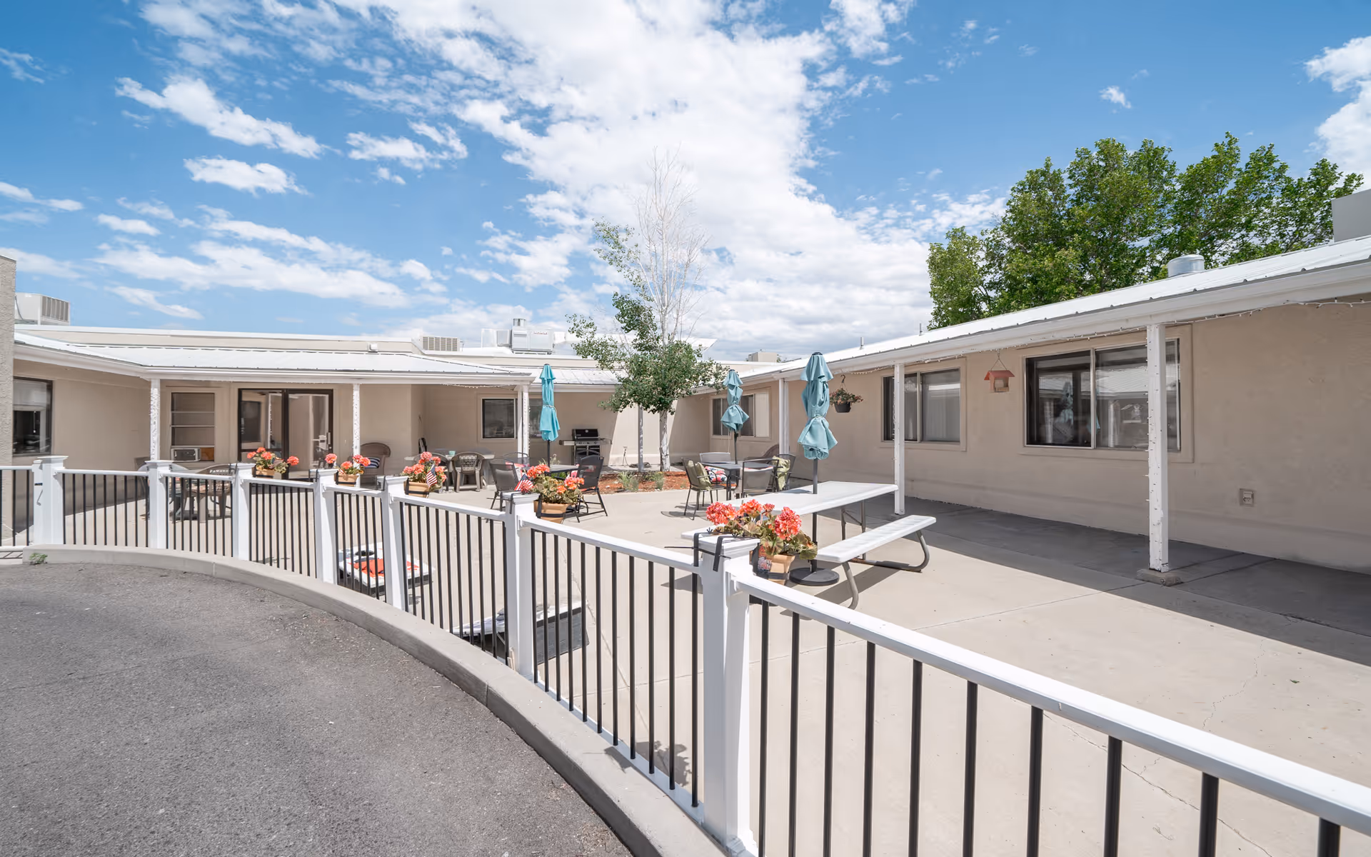 Outdoor courtyard area of Mantey Heights Rehabilitation and Care Center with a curved white and black railing, several tables with chairs and closed umbrellas, potted flowers on the railing, and a few trees under a partly cloudy blue sky.