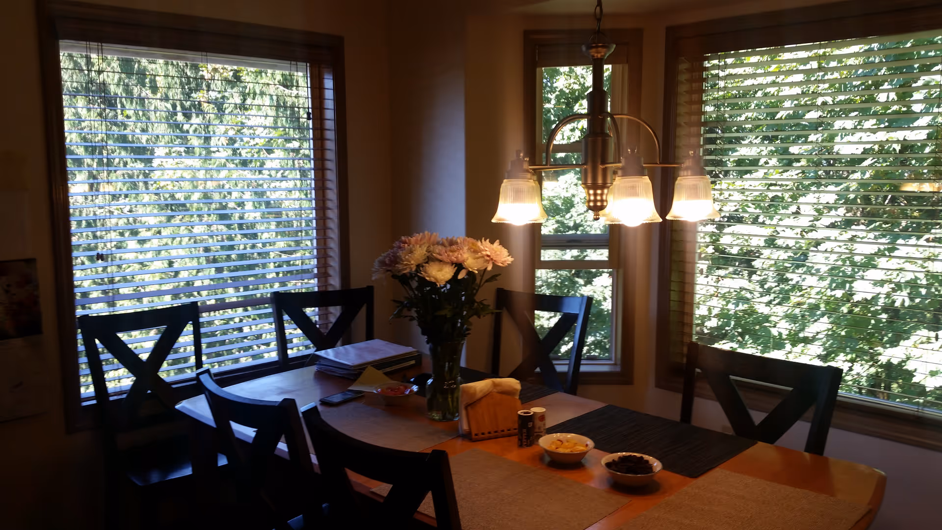 Dining area with a wooden table surrounded by six black chairs. The table has a vase with flowers, some papers, a tissue box, and two bowls with snacks. Large windows with wooden blinds let in natural light and show green foliage outside. A ceiling light fixture with four lamps hangs above the table.