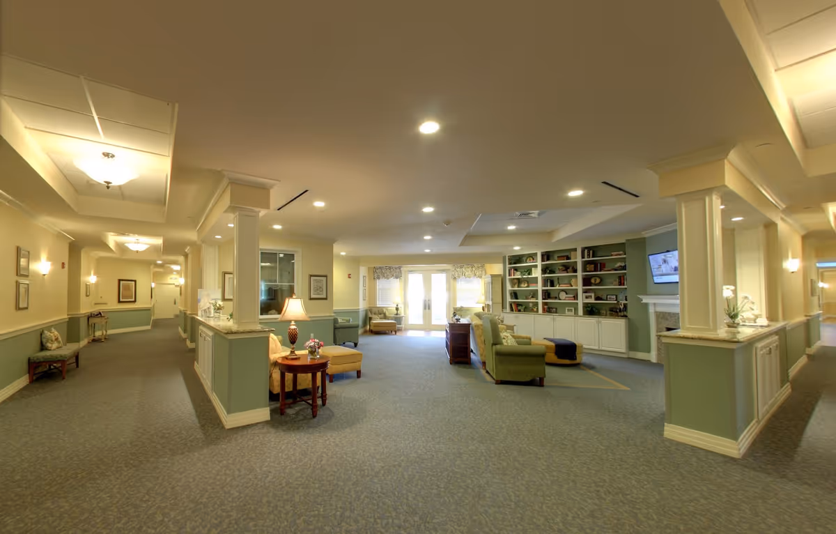 A spacious and well-lit common area in a senior living facility with comfortable seating including armchairs and ottomans, a fireplace with a mounted TV above it, built-in shelves with decorative items, and a long hallway extending on both sides. The walls are painted in soft green and cream tones, and the ceiling has recessed lighting and decorative light fixtures.