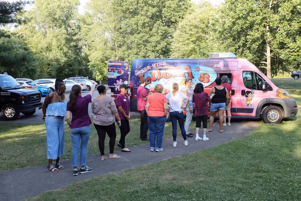 A group of people standing in line outdoors on a paved path, waiting to be served from a colorful pink and purple food truck with the name 'Lucca's' on it. The scene is set in a grassy area with trees and parked cars in the background.