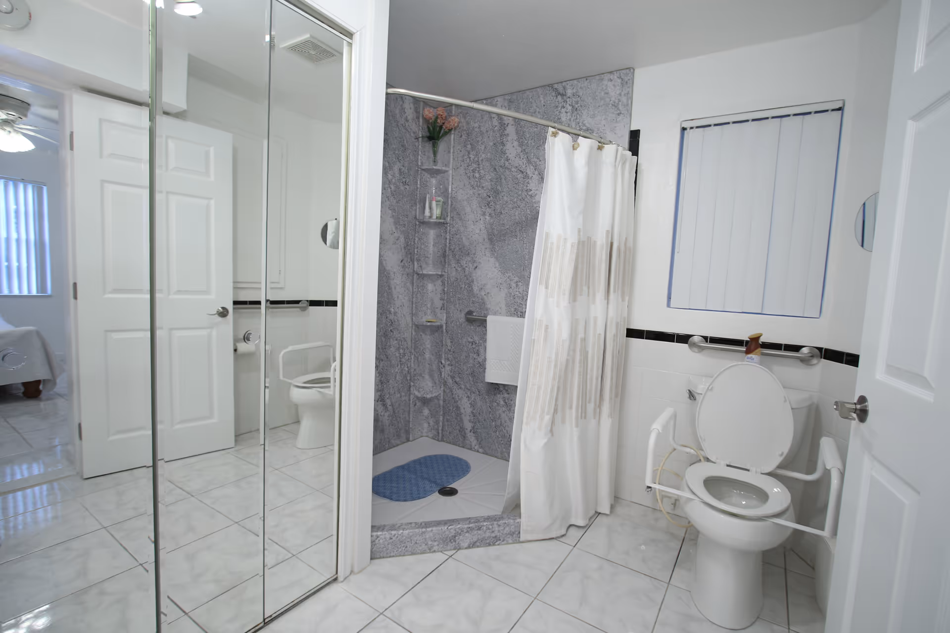 Accessible bathroom with a walk-in shower, shower curtain, raised toilet with arm rails and grab bars, and mirrored closet doors.