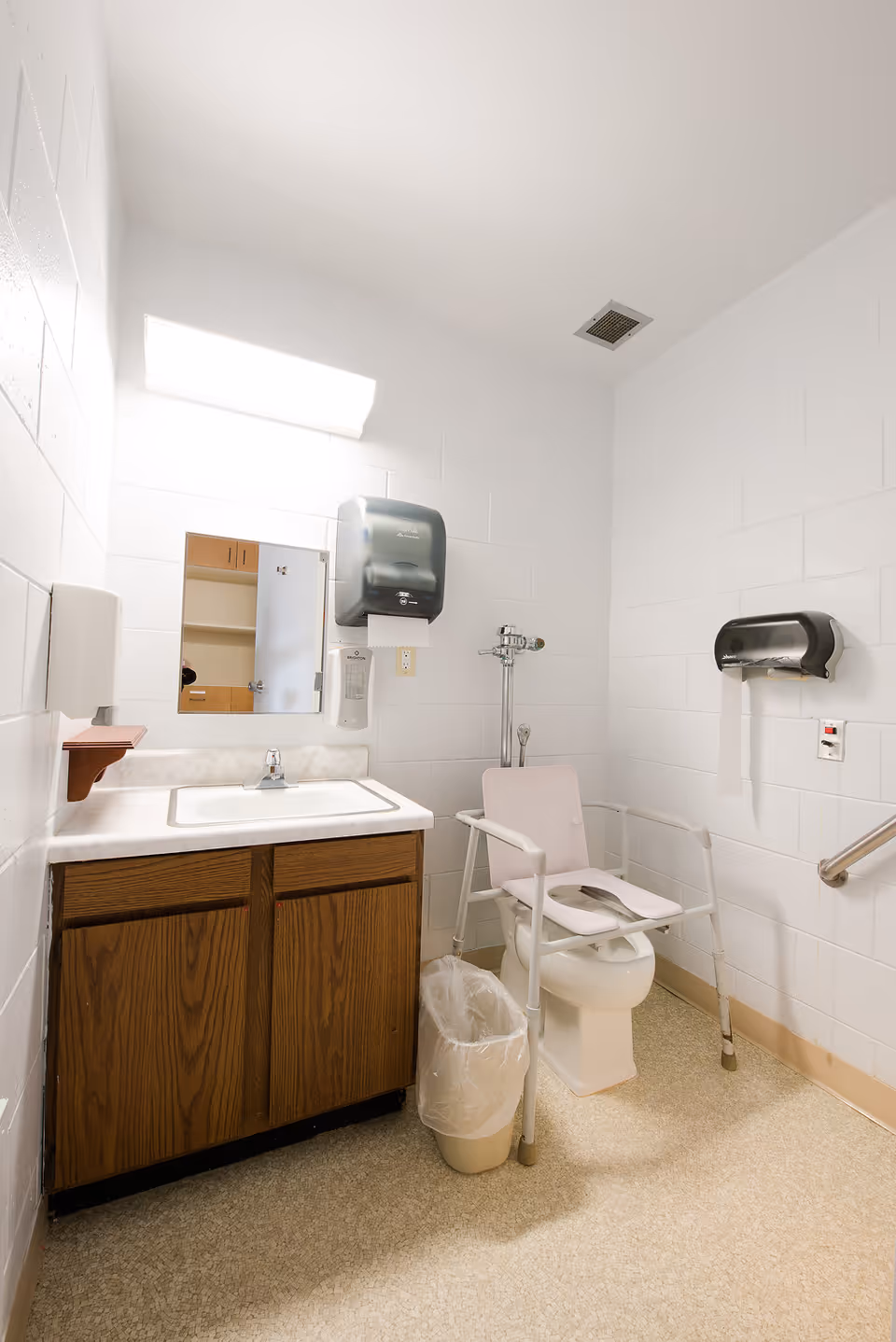 A small, accessible bathroom with white walls and beige flooring. It features a wooden cabinet with a white sink and a mirror above it. There is a wall-mounted paper towel dispenser and a soap dispenser next to the sink. The toilet has a raised seat with armrests for accessibility, and there is a trash can with a plastic liner next to the cabinet. A handrail is mounted on the wall near the toilet.