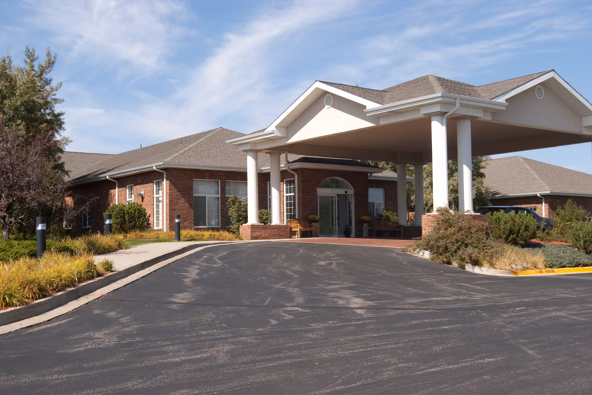Exterior view of a single-story brick building with a covered entrance supported by white columns, surrounded by landscaped bushes and trees under a partly cloudy sky.