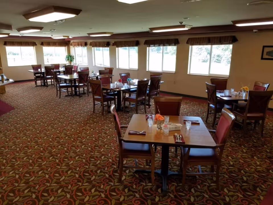 A dining room in a senior living facility with multiple wooden tables and chairs arranged neatly. Each table is set with glasses, napkins, and silverware, and some have small flower arrangements. Large windows along the wall let in natural light, and the room has patterned carpet and ceiling lights.